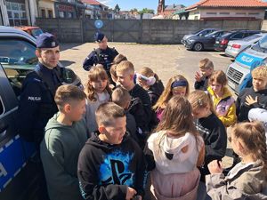 na zdjęciu widać stojących przy radiowozie dwóch policjantów i gromadkę dzieci uczniów szkoły podstawowej