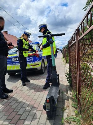 na pierwszym planie zdjęcia widać hulajnogę elektryczną którą kierujący stojący tuż obok niej zaparkował na chodniku w tle dwie policjantki które dokonują legitymowania mężczyzny a dalej w tle policyjny radiowóz