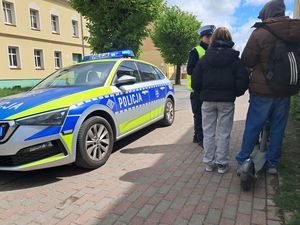 na zdjęciu widać zaparkowany na krawędzi chodnika radiowóz obok niego policjantka która rozmawia z dwójką młodzieży która jechała na jednej hulajnodze