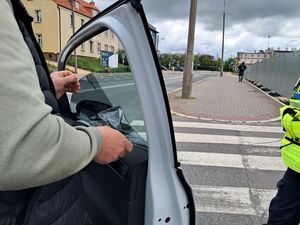 na zdjęciu widać fragment kamizelki policyjnej od stojącej poza kadrem policjantki przy otwartych drzwiach kontrolowanego samochodu stoi kierowca który odrywa folię przyciemniającą przednią boczną szybę w samochodzie