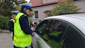 na zdjęciu widać policjantkę stojącą przy otwartym oknie kontrolowanego samochodu która pokazuje kontrolowanemu kierowcy urządzenie do mierzenia przejrzystości szyb z wynikiem pomiaru