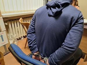 na zdjęciu widać mężczyzn siedzącego na przesłuchaniu z zakutymi w kajdanki ręce na tyle pleców