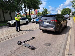na zdjęciu widać dwupasmową jezdnię na na których widać samochody po środku na wysepce stoi radiowóz obok niego policjant który coś pisze a obok niego na ulicy leży hulajnoga