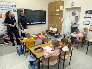 na zdjęciu widać policjantki stojące obok tablicy w klasie widać dzieci siedzące w ławkach i nauczycielkę stojącą w pobliżu drzwi
