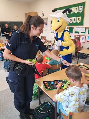 na zdjęciu widać policjantkę i dzieci w klasie w tle osoba przebrana w maskotkę mrówki rozdającą odblaski