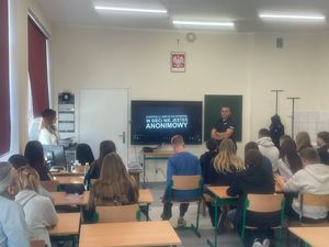 na zdjęciu widać grupę siedzącej młodzieży przed tablicą stoi policjant i kurator sądowa a ekranie wyświetlana jest prezentacja