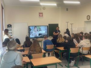 na zdjęciu widać grupę siedzącej młodzieży przed tablicą stoi policjant i kurator sądowa a ekranie wyświetlana jest prezentacja