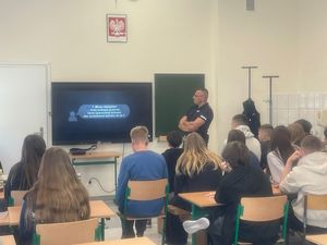 na zdjęciu widać grupę siedzącej młodzieży przed tablicą stoi policjant na ekranie wyświetlana jest prezentacja
