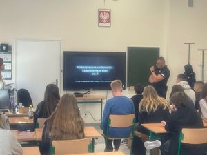 na zdjęciu widać grupę siedzącej młodzieży przed tablicą stoi policjant i kurator sądowa na ekranie wyświetlana jest prezentacja