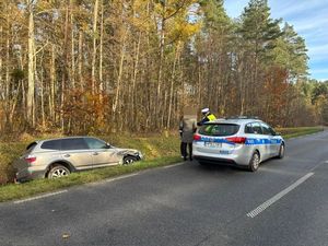 na zdjęciu widać w scenerii leśnej stojący na jezdni radiowóz obok niego w przydrożnym rowie samochód osobowy który był uczestnikiem kolizji przy radiowozie stoi policjant i dwie osoby