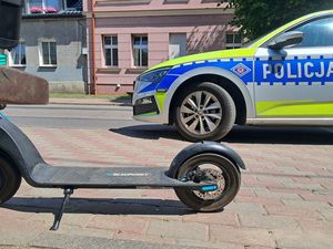 na zdjęciu widać zaparkowaną na chodniku hulajnogę obok niej radiowóz w tle widać ulicę za nią kamiennicę
