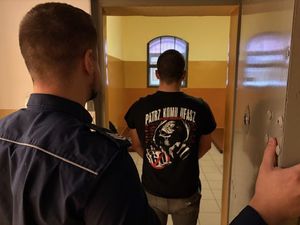 na zdjęciu widać policjanta który otworzył drzwi do celi a wewnątrz niej widać stojącego tyłem mężczyznę który ma na sobie koszulkę z napisem patrz komu ufasz