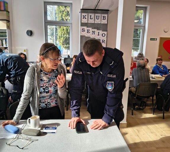 na zdjęciu widać policjanta który pokazuje seniorce jak na telefonie komórkowym obsługiwać krajową mapę zagrożeń bezpieczeństwa