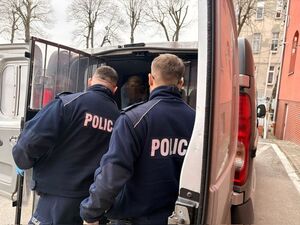 na zdjęciu widać dwóch policjantów stojących na tyle radiowozu busa nieoznakowanego należącego do konwojówki widać jak w aucie siedzi zatrzymany mężczyzna otwarte są okratowane drzwi