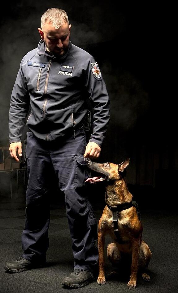 na zdjęciu widać umundurowanego policjanta ciemne tło w koło obok policjanta siedzi pies i obaj patrzą na siebie