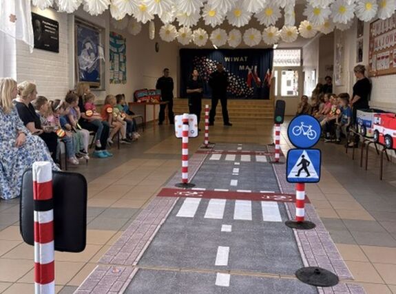 na zdjęciu widać rozłożone na holu szkoły zabawkowe miasteczko ruchu drogowego wkoło na ławkach siedzą dzieci i nauczyciele z przodu widać policjantów