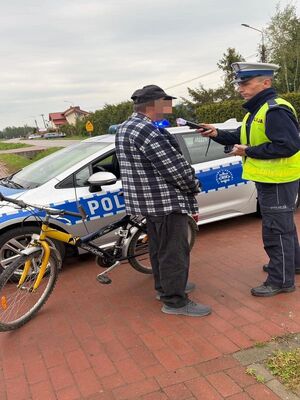 na zdjęciu z prawej strony stoi policjant który trzyma w ręku alkomat i bada nim rowerzystę w koszuli w kratę  ciemnych spodniach oraz czapce z daszkiem który stoi obok w tle jest radiowóz i żółtoczarny rower