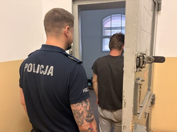 na zdjęciu widać policjanta umundurowanego który stoi w drzwiach do cli a przed nim tyłem osoba zatrzymana osadzona w tym pomieszczeniu