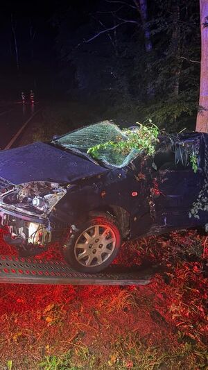 na zdjęciu widać porozbijany samochód który jest wyciągany z rowu przez holownik w tle widać las i dwóch funkcjonariuszy straży pożarnej