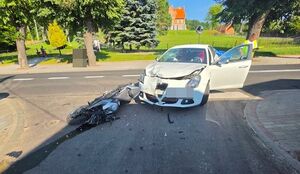 na zdjęciu widać skrzyżowanie ulic przewrócony na jezdni motocykl obok niego białe auto osobowe w tle widać kościół na górce a obok siedzące na ławce przy chodniku osoby